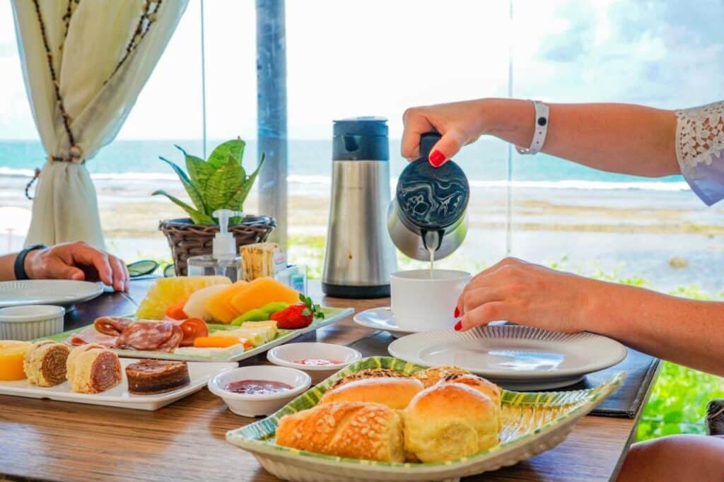 Uma pessoa serve chá em uma mesa com pão, frutas, queijo e bolos no Summerville Resort - All Inclusive. Vista do oceano ao fundo, com uma planta próxima.