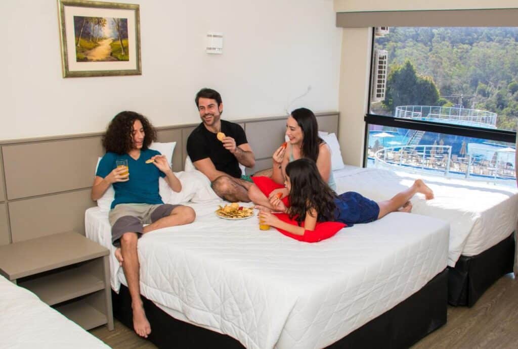 Quatro pessoas sentam-se em uma cama em um quarto no Cassino All Inclusive Resort Poços de Caldas compartilhando lanches. O quarto tem uma grande janela com vista para árvores e uma piscina.