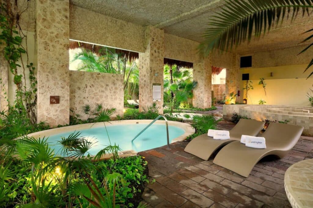 Spa interno do Grand Palladium Colonial Resort & Spa com uma pequena piscina circular cercada por vegetação, duas espreguiçadeiras bege e uma parede de pedra texturizada.