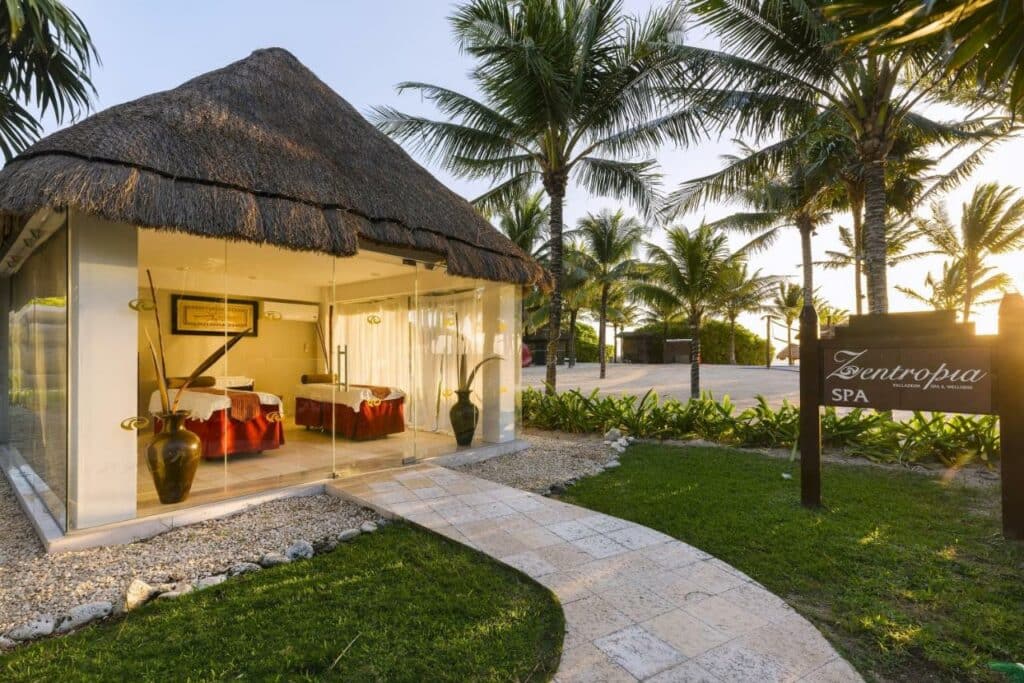 Um spa com paredes de vidro e telhado de palha no Grand Palladium Colonial Resort & Spa, cercado por palmeiras, com uma placa dizendo "Zentropia Spa" ao pôr do sol.