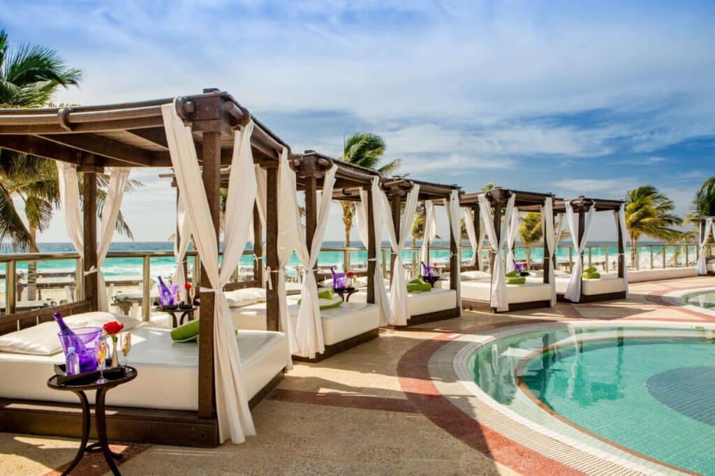 Cabanas à beira da piscina do Hyatt Zilara Cancun com cortinas brancas e espreguiçadeiras ao lado de uma piscina circular com vista para o mar e palmeiras ao fundo. Esse é um dos melhores resorts all inclusive no Caribe.