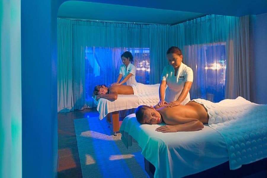 Duas pessoas recebendo massagens em mesas em uma sala com iluminação azul no Renaissance Wind Creek Aruba Resort. Dois terapeutas estão de pé ao lado delas.