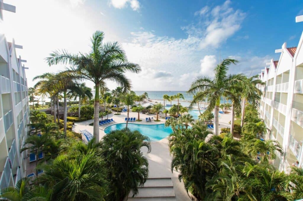 Uma vista do Renaissance Wind Creek Aruba Resort com palmeiras, uma piscina cheia de curvas cercada por espreguiçadeiras e edifícios em ambos os lados que levam ao oceano. Esse é um dos melhores resorts all inclusive no Caribe.