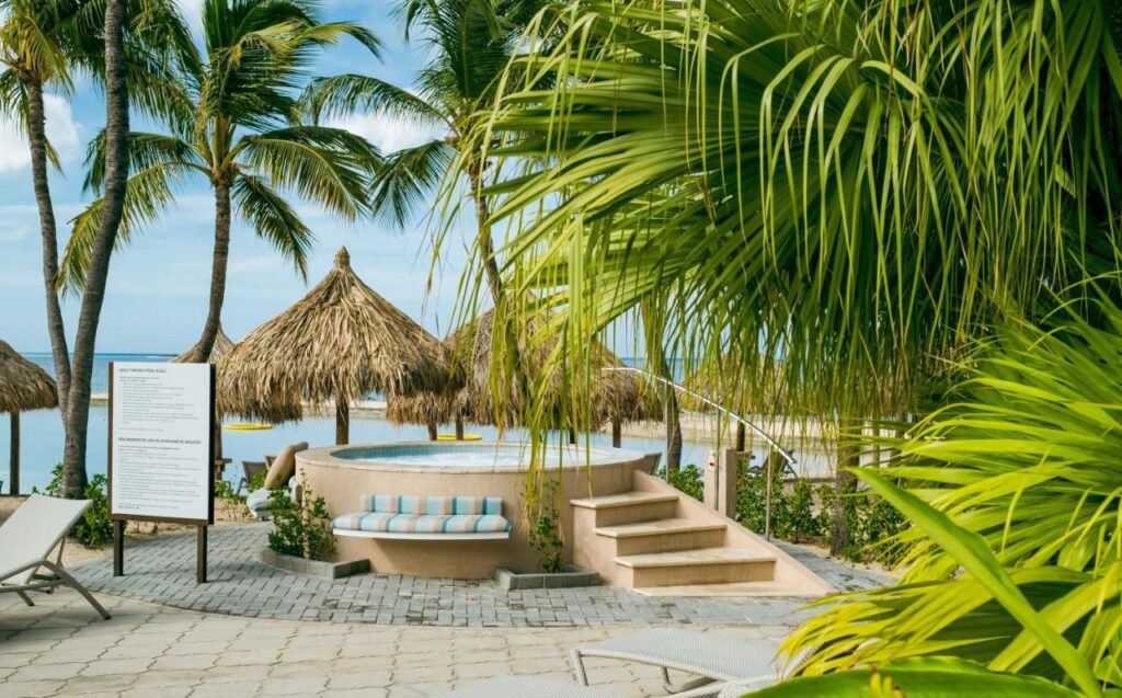 Um cenário de praia tropical com palmeiras, guarda-sóis de palha, um banco curvo e uma pequena piscina elevada perto do oceano no Renaissance Wind Creek Aruba Resort.