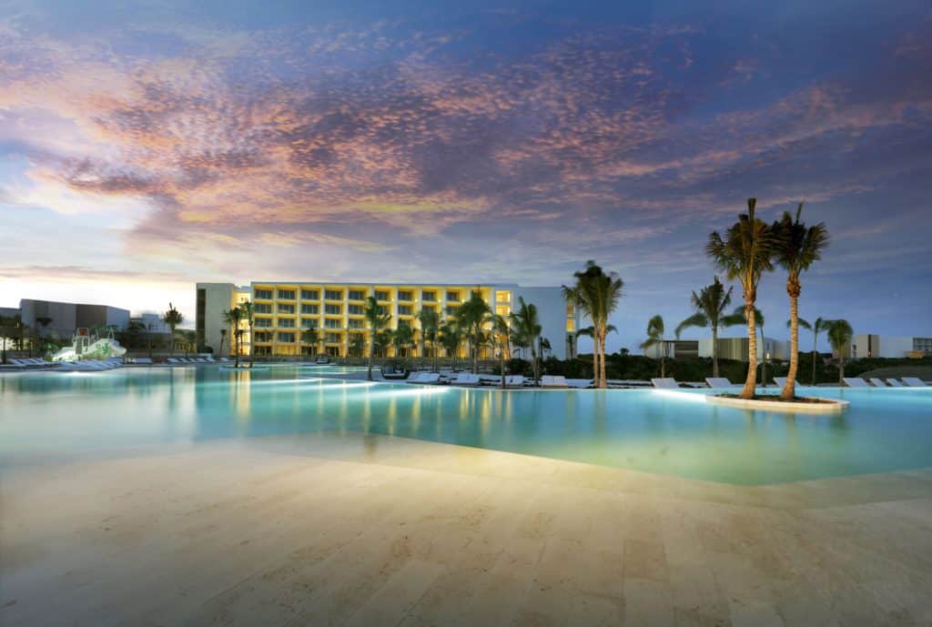 Uma grande piscina ao ar livre refletindo as cores do pôr do sol, com palmeiras e o Grand Palladium Select Costa Mujeres ao fundo. Esse é um dos melhores resorts all inclusive no Caribe.