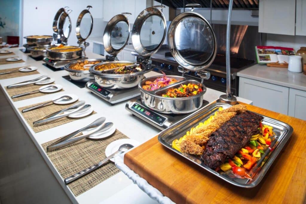 Um bufê no S Hotel Montego Baycom vários pratos quentes em rechauds, incluindo carne assada, vegetais, arroz e macarrão, em um balcão. Esse é um dos melhores resorts all inclusive no Caribe.