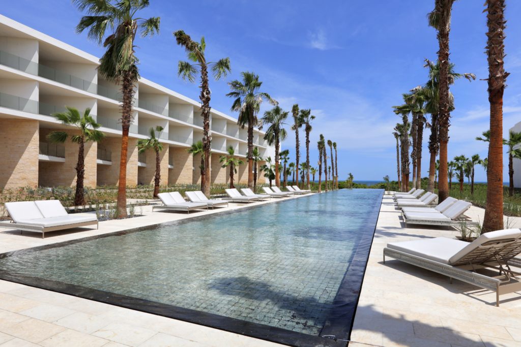 Uma longa piscina ao ar livre ladeada por palmeiras e espreguiçadeiras brancas. Um edifício moderno do Grand Palladium Select Costa Mujeres está ao fundo sob um céu azul claro. Esse é um dos melhores resorts all inclusive no Caribe.