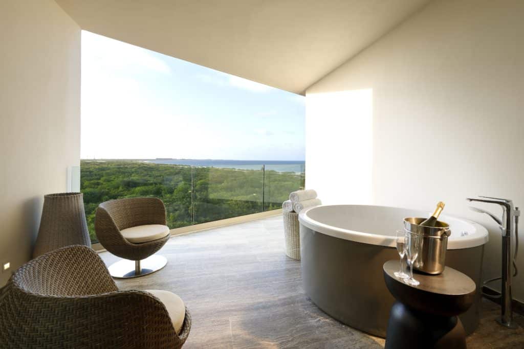 Banheiro luxuoso do Grand Palladium Select Costa Mujeres com banheira redonda, cadeiras de vime, champanhe no gelo e vista panorâmica da vegetação através de grandes janelas de vidro.