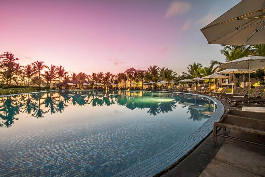 Grande piscina ao ar livre no Hard Rock Hotel & Casino Punta Cana reflete palmeiras e um céu de pôr do sol. Cadeiras de estar e guarda-sóis alinham-se à beira da piscina, com um prédio ao fundo. Esse é um dos melhores resorts all inclusive no Caribe.