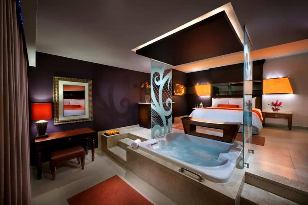 Quarto moderno do Hard Rock Hotel & Casino Punta Cana com uma cama grande, uma escrivaninha e uma banheira aberta separada por uma divisória de vidro. Iluminação e decoração aconchegantes. Esse é um dos melhores resorts all inclusive no Caribe.
