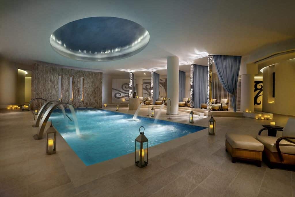 Luxuosa área de piscina coberta no Hard Rock Hotel & Casino Punta Cana com espreguiçadeiras, lanternas acesas, cascatas e uma grande claraboia circular.