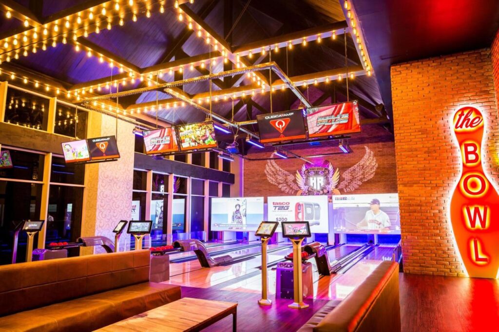 Uma pista de boliche iluminada no Hard Rock Hotel & Casino Punta Cana com várias pistas, TVs suspensas, letreiros luminosos e uma área de estar com sofás.