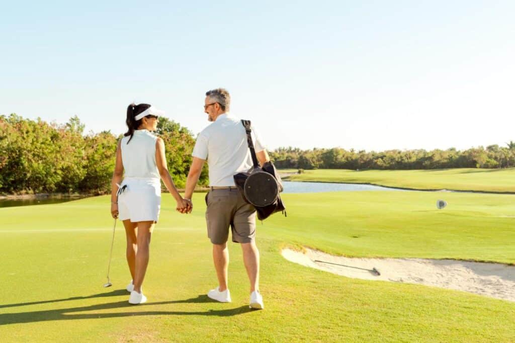 Um casal de mãos dadas caminhando em um campo de golfe ensolarado no Hard Rock Hotel & Casino Punta Cana, com tacos de golfe e vestindo trajes casuais de golfe.