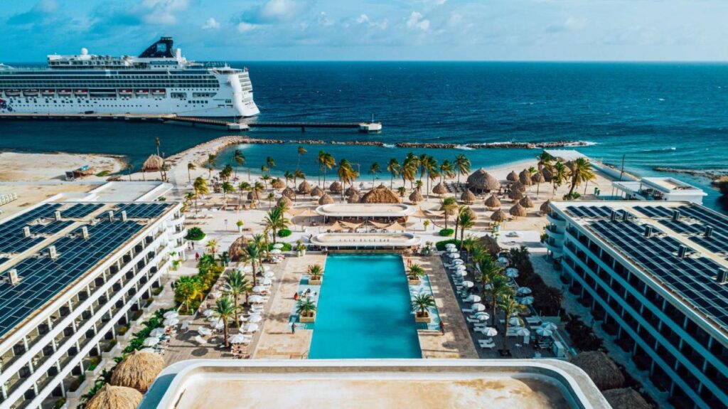Vista aérea do Mangrove Beach Corendon com uma grande piscina, palmeiras, edifícios ao redor e um navio de cruzeiro ao fundo. Esse é um dos melhores resorts all inclusive no Caribe.
