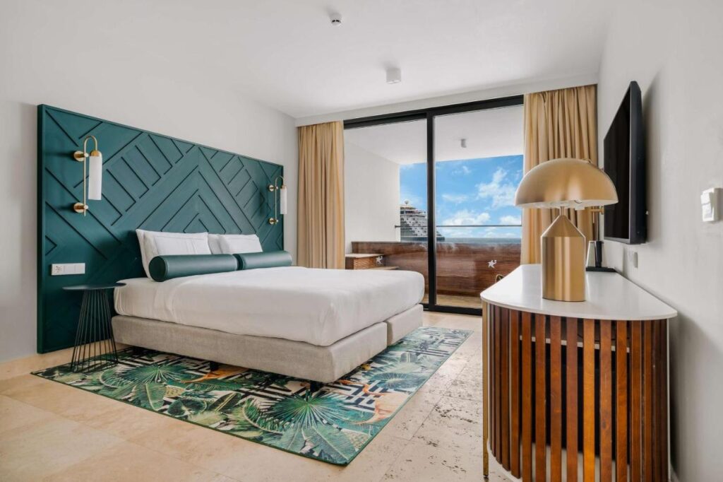 Quarto moderno do Mangrove Beach Corendon com uma cama grande, parede com detalhes em verde, tapete estampado e vista da sacada. Uma luminária e uma TV ficam em um armário de madeira.