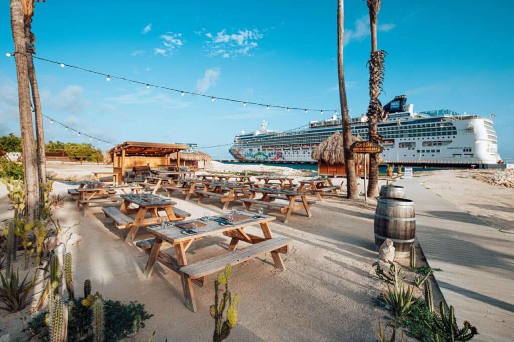 Um restaurante à beira-mar no Mangrove Beach Corendon com mesas de madeira, luzes de corda e um grande navio de cruzeiro atracado nas proximidades sob um céu azul claro.