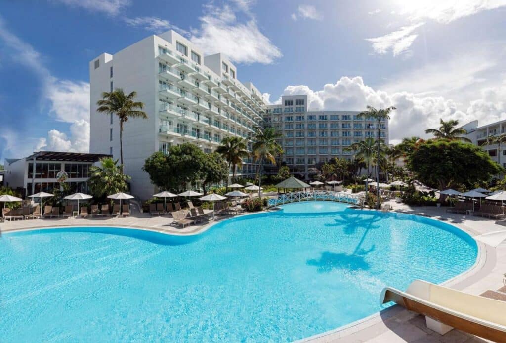 Uma grande piscina com espreguiçadeiras e guarda-sóis é cercada pelo Sonesta Maho Beach Resort de vários andares e palmeiras sob um céu parcialmente nublado. Esse é um dos melhores resorts all inclusive no Caribe.