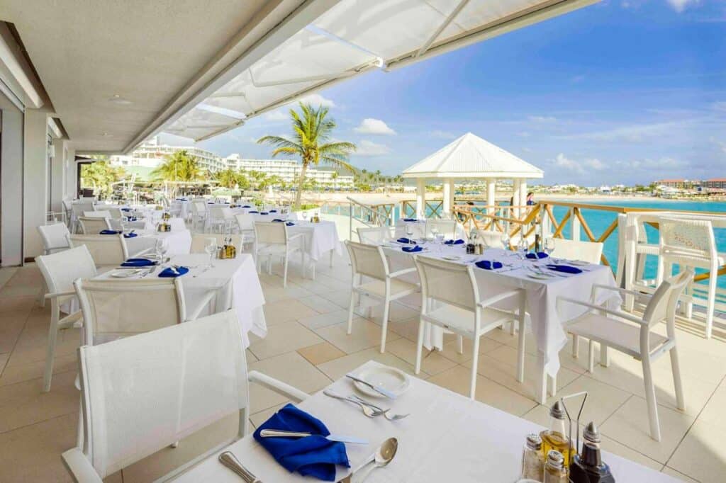 Restaurante ao ar livre à beira-mar no Sonesta Maho Beach Resort com mesas e cadeiras brancas, mesa com guardanapos e copos azuis, com vista para o oceano e o gazebo. Esse é um dos melhores resorts all inclusive no Caribe.