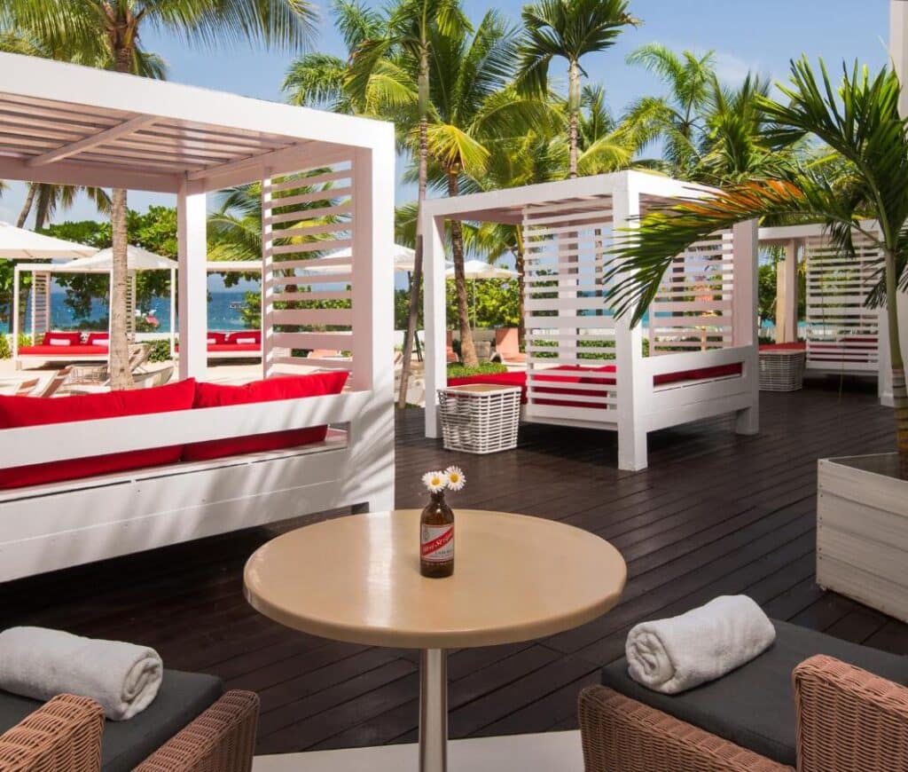 Área de estar ao ar livre no S Hotel Montego Bay com cabanas brancas, almofadas vermelhas, palmeiras e uma mesa redonda com uma garrafa de bebida e uma flor em cima. Esse é um dos melhores resorts all inclusive no Caribe.