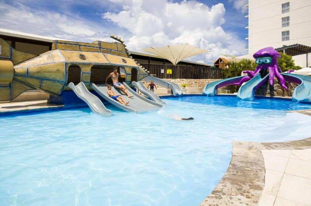 Crianças aproveitando toboáguas ao lado de uma estrutura de polvo roxo em uma piscina sob um céu parcialmente nublado no Sonesta Maho Beach Resort. Esse é um dos melhores resorts all inclusive no Caribe.