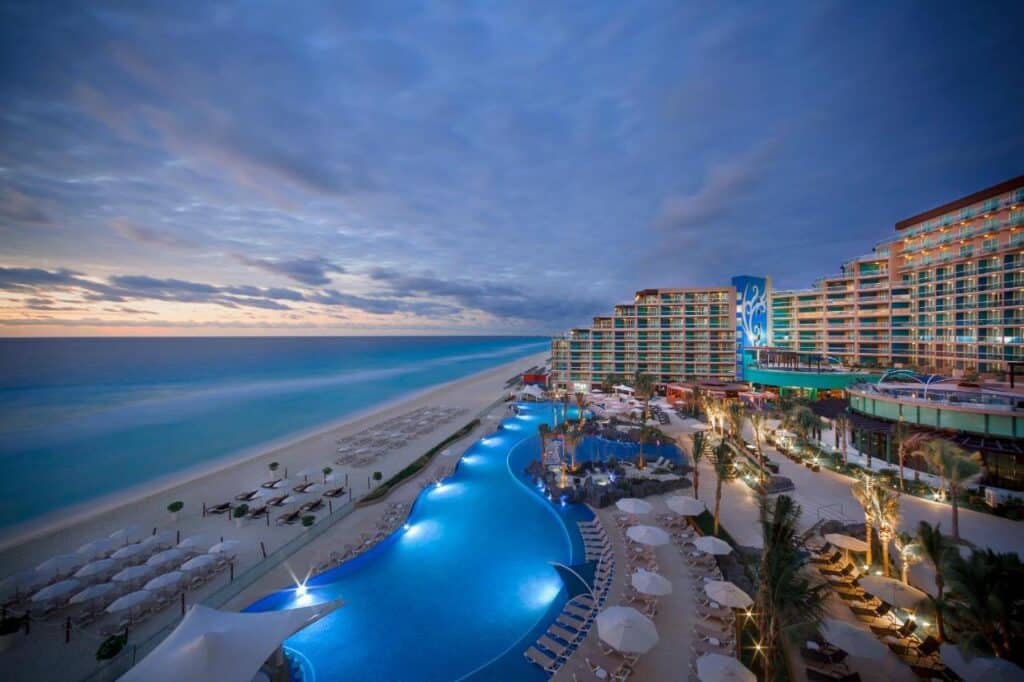 Vista aérea do Hard Rock Hotel Cancun à beira-mar ao anoitecer, com uma grande piscina, fileiras de cadeiras de praia e ondas do mar sob um céu nublado. Esse é um dos melhores resorts all inclusive no Caribe.
