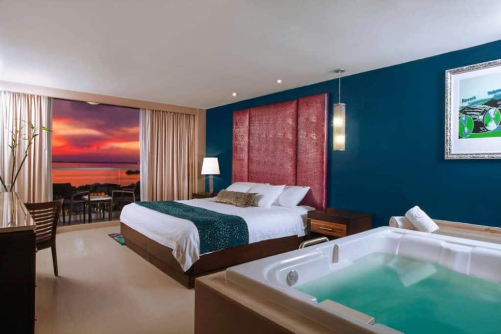Quarto do Hard Rock Hotel Cancun com cama king-size, jacuzzi interna, escrivaninha e varanda com vista para o pôr do sol no mar.