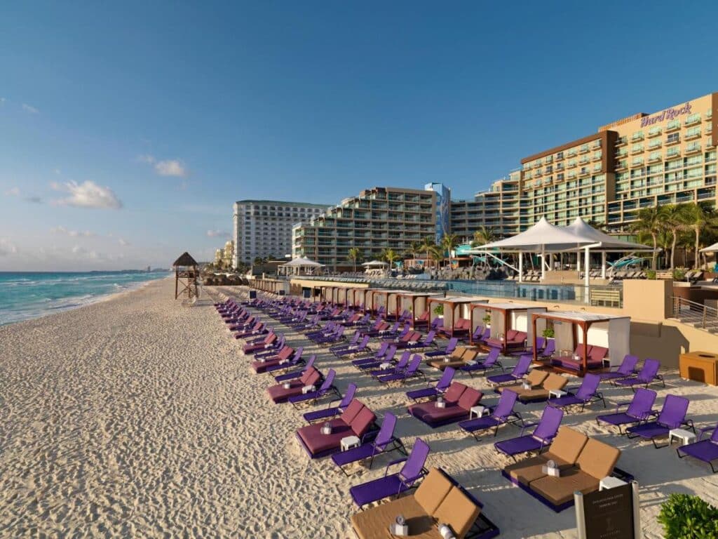 O Hard Rock Hotel Cancun à beira-mar com fileiras de espreguiçadeiras roxas e cabanas brancas na praia arenosa sob um céu limpo perto do oceano. Esse é um dos melhores resorts all inclusive no Caribe.