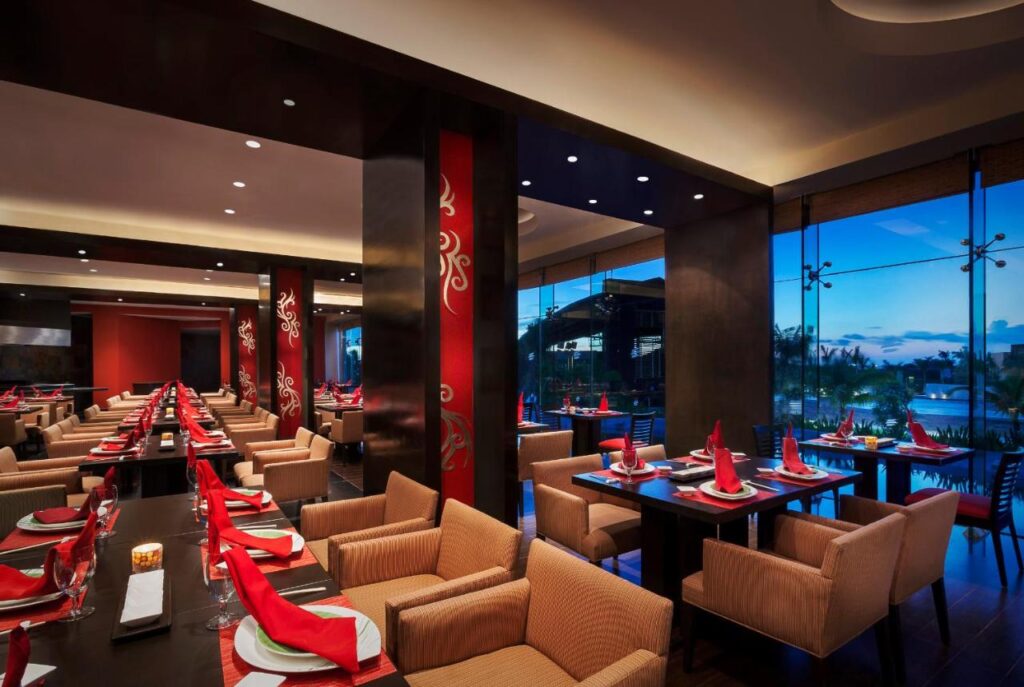 Um restaurante moderno do Hard Rock Hotel Cancun com mesas bem dispostas, guardanapos vermelhos, cadeiras bege e grandes janelas com vista para o fim da tarde. Esse é um dos melhores resorts all inclusive no Caribe.