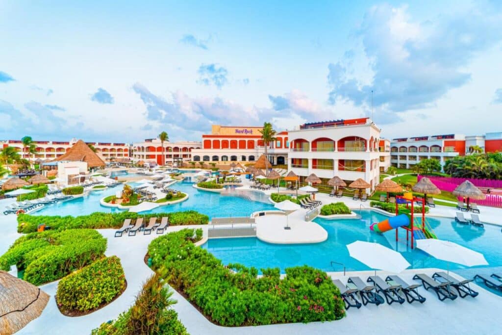 O Hard Rock Hotel Riviera Maya com várias piscinas, espreguiçadeiras e estruturas coloridas, tendo como pano de fundo céu azul e nuvens dispersas. Esse é um dos melhores resorts all inclusive no Caribe.