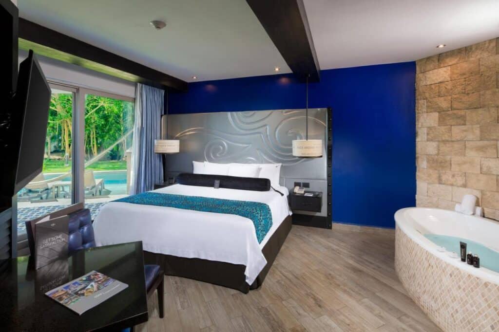 Quarto moderno do Hard Rock Hotel Riviera Maya com cama grande, TV de tela plana e jacuzzi. Há vista para uma piscina e vegetação através de portas de vidro deslizantes.