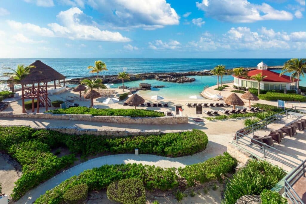 Vista panorâmica do Hard Rock Hotel Riviera Maya de praia com espreguiçadeiras, um mirante, palmeiras e um oceano azul claro sob um céu parcialmente nublado. Esse é um dos melhores resorts all inclusive no Caribe.