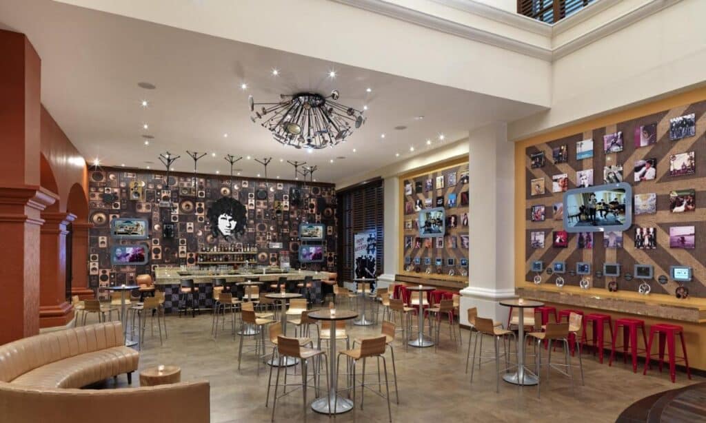 Um café moderno do Hard Rock Hotel Riviera Maya com mesas e cadeiras de madeira, paredes decoradas com fotos e obras de arte e um lustre diferenciado no alto.