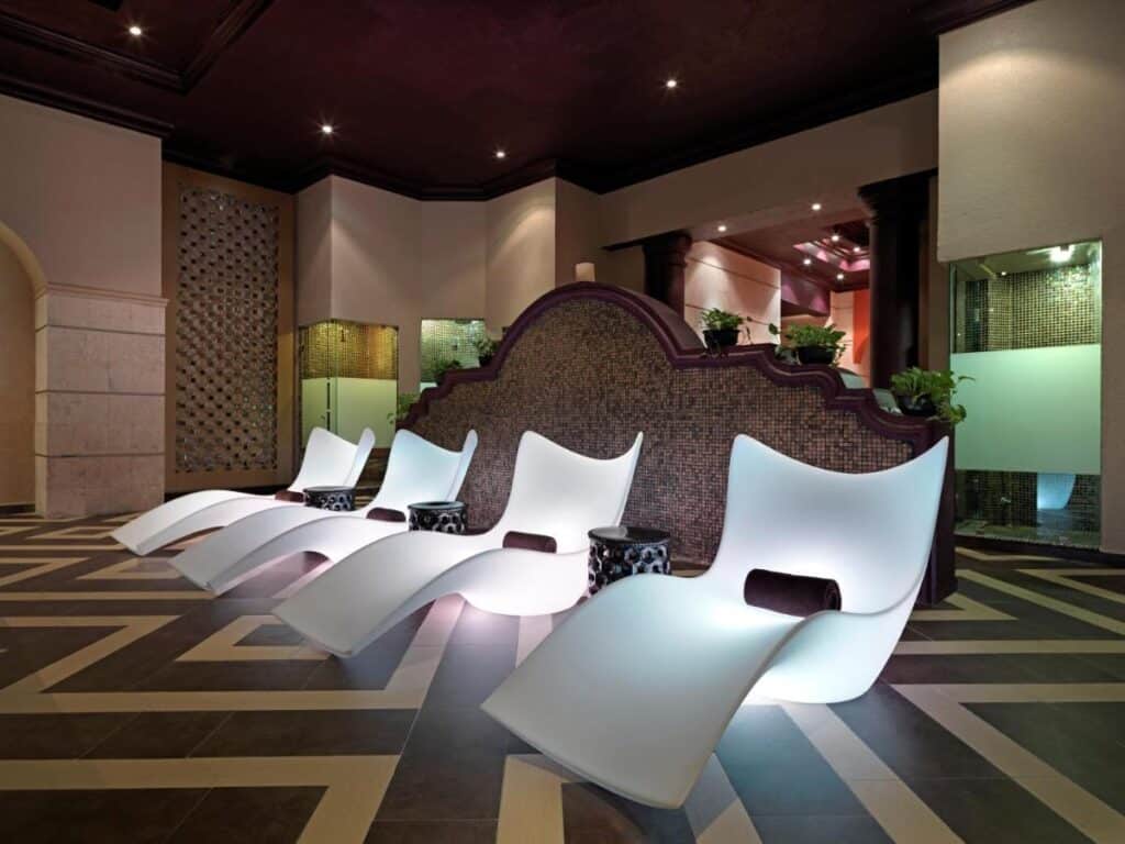 Sala moderna do Hard Rock Hotel Riviera Maya com cadeiras brancas curvas, pequenas mesas laterais e uma parede decorativa com plantas, dispostas em um piso geométrico.