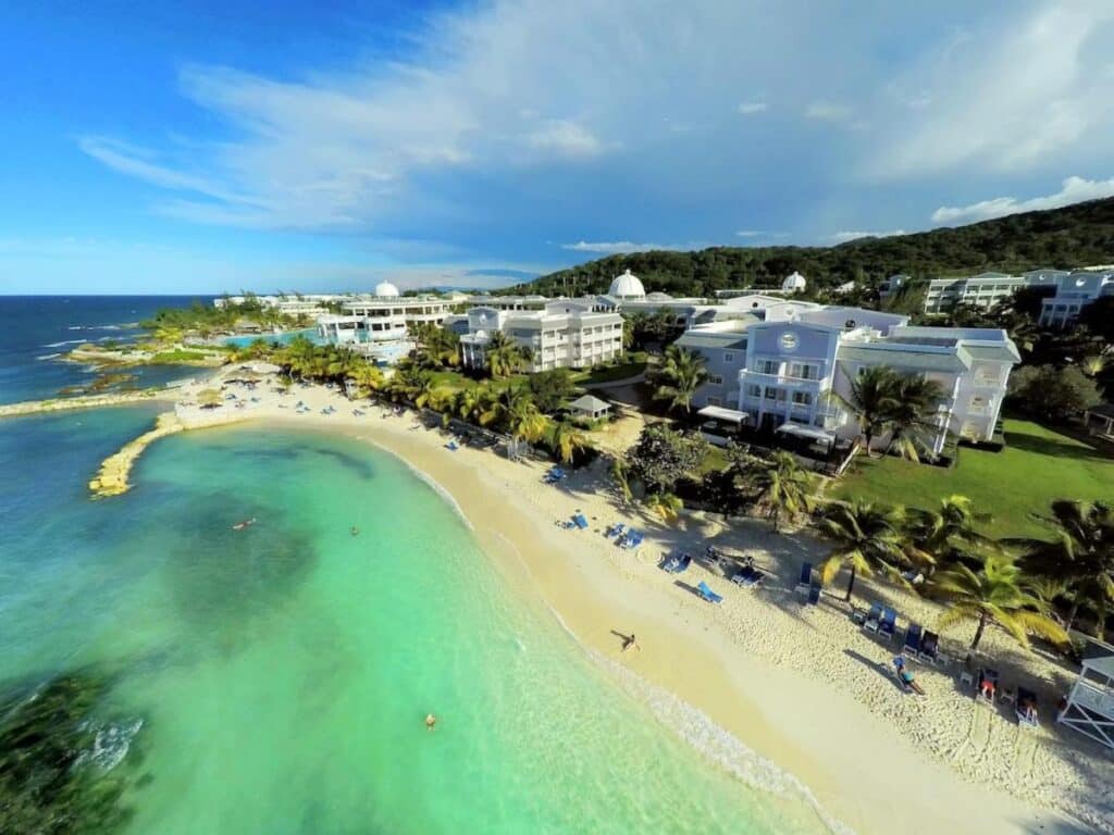 Vista aérea do Grand Palladium Jamaica Resort & Spa à beira-mar com edifícios brancos, palmeiras e águas azul-turquesa sob um céu parcialmente nublado. Esse é um dos melhores resorts all inclusive no Caribe.