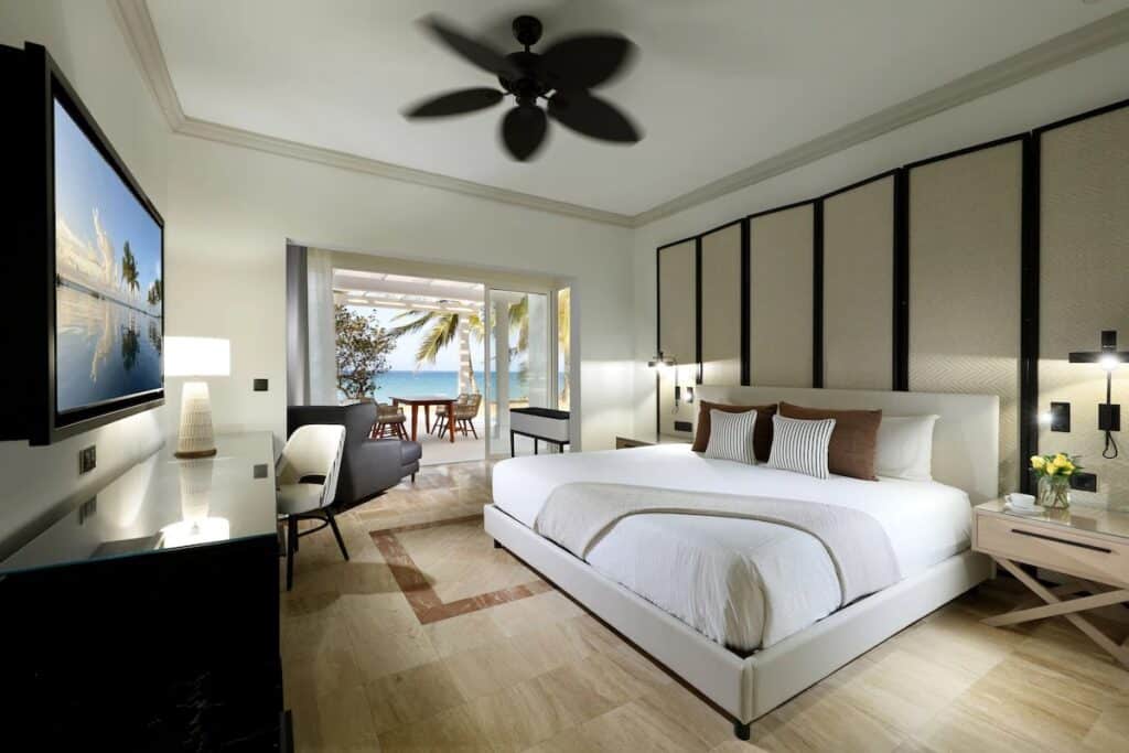 Um quarto moderno do Grand Palladium Jamaica Resort & Spa com uma cama grande, poltrona e uma TV montada na parede. Uma porta de vidro abre para um pátio com vista para o oceano.