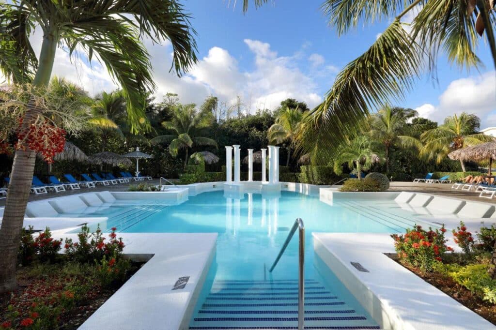 Uma piscina do Grand Palladium Jamaica Resort & Spa com pilares em uma extremidade, cercada por espreguiçadeiras, palmeiras e vegetação exuberante sob um céu azul. Esse é um dos melhores resorts all inclusive no Caribe.