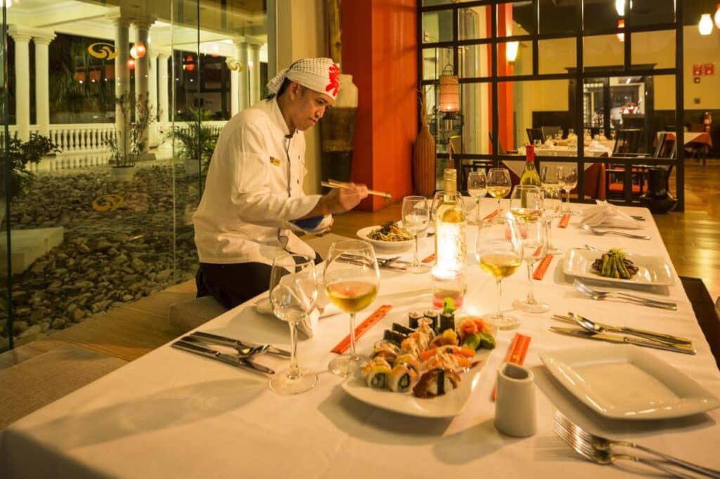 Chef preparando pratos em uma mesa de restaurante do Grand Palladium Jamaica Resort & Spa com sushi, taças de vinho e velas, em uma área de jantar bem iluminada.