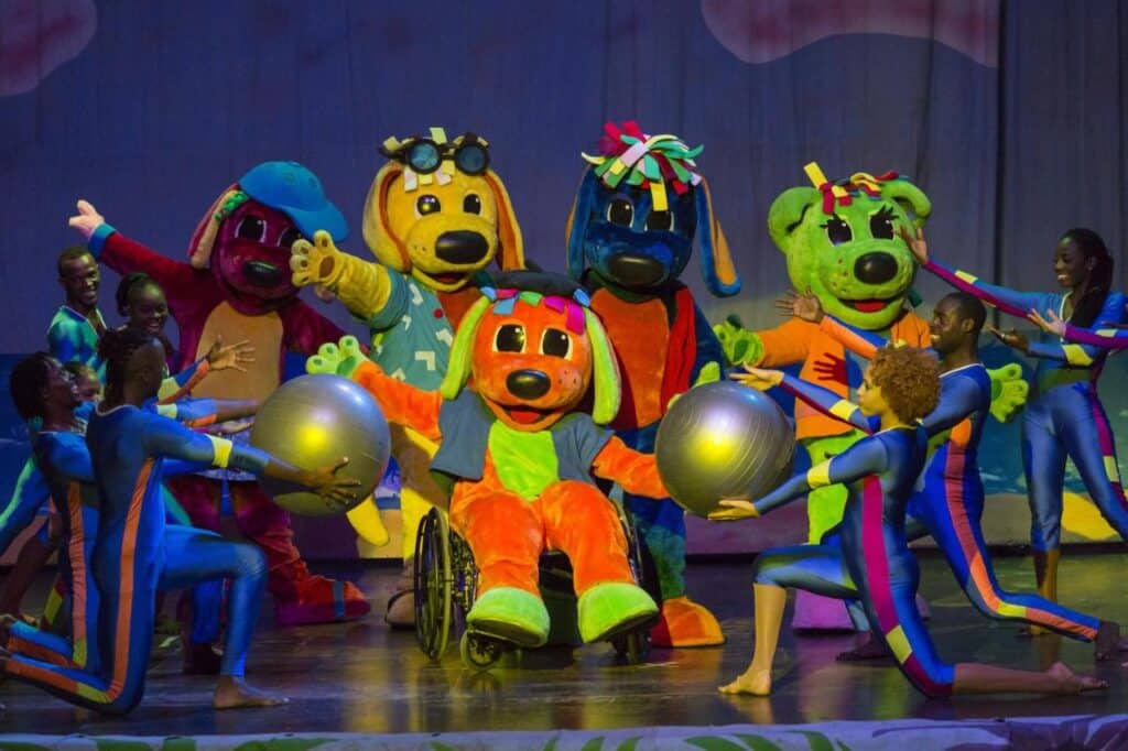 Artistas vestidos com fantasias coloridas de cachorro e macacões dançam no palco do Grand Palladium Jamaica Resort & Spa, dois deles segurando grandes bolas prateadas.