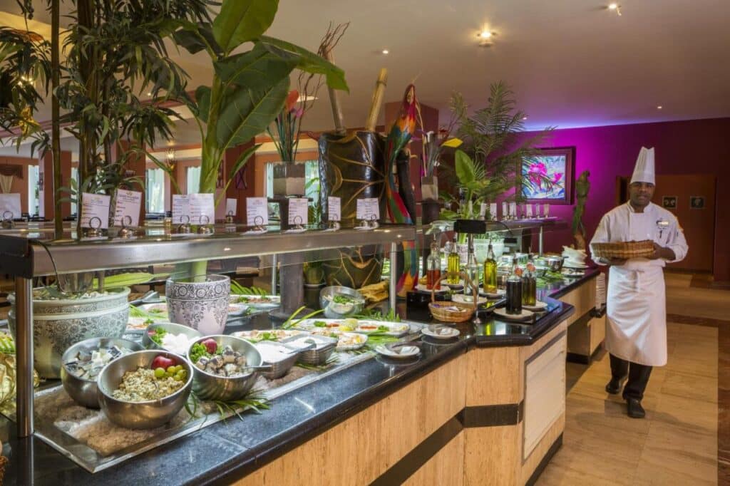 Um chef de uniforme branco e chapéu alto está perto de um bufê com vários pratos, plantas e garrafas em um restaurante bem iluminado no Grand Palladium Jamaica Resort & Spa. Esse é um dos melhores resorts all inclusive no Caribe.