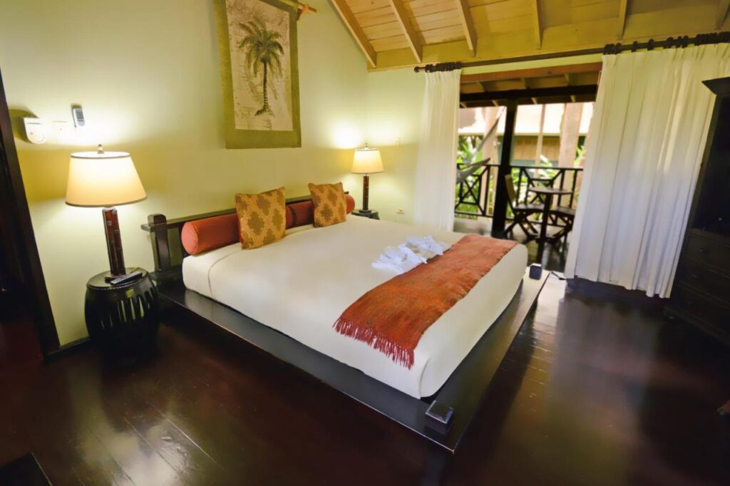 Um quarto aconchegante do Sunset at the Palms Resort com uma cama grande, teto de madeira, luminárias de cabeceira e vista para uma varanda com cadeiras e uma mesa do lado de fora.