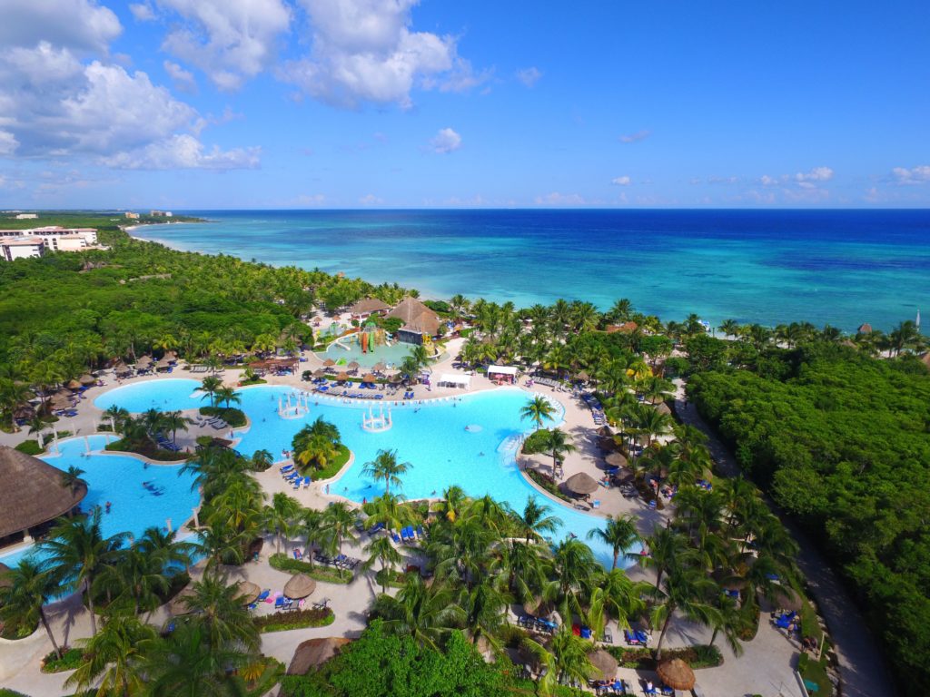 Vista aérea do Grand Palladium Kantenah Resort & Spa com grandes piscinas azuis cercadas por árvores, perto de uma praia com águas cristalinas azul-turquesa. Esse é um dos melhores resorts all inclusive no Caribe.