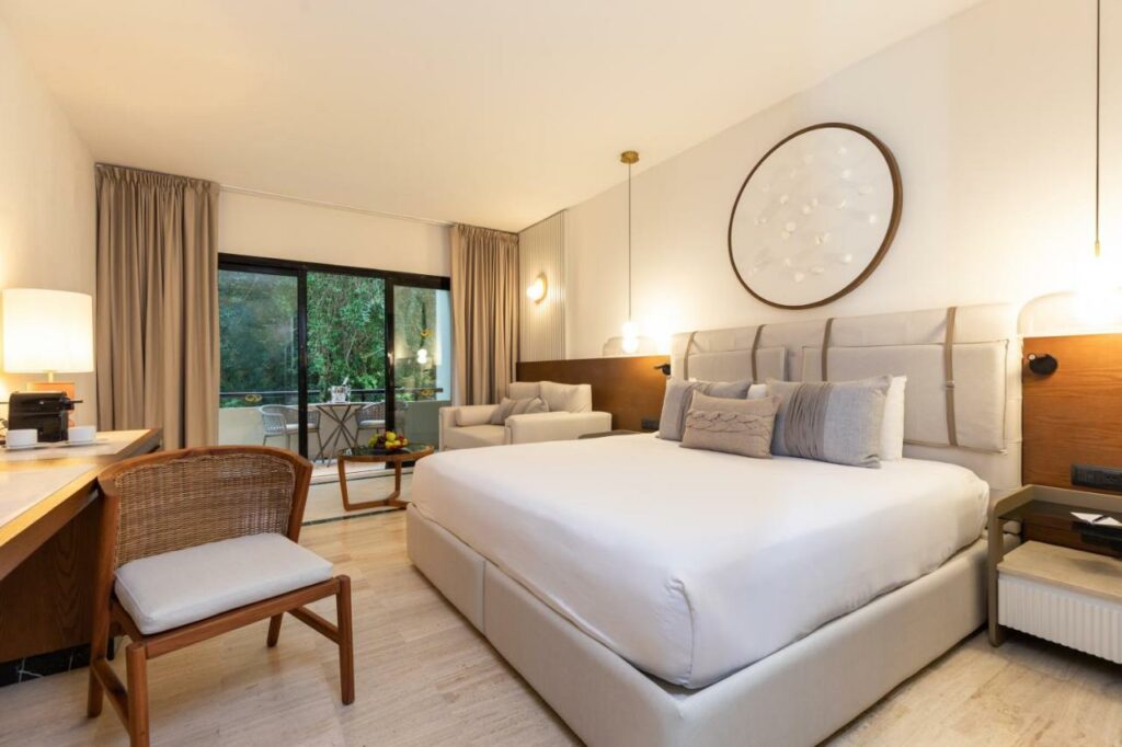 Um quarto moderno do Grand Palladium Kantenah Resort & Spa com uma cama grande, área de estar, escrivaninha e varanda com vista para a área verde do lado de fora.