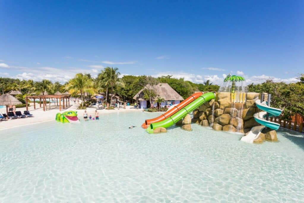 Piscina infantil do Grand Palladium Kantenah Resort & Spa com toboáguas e um espelho d'água em formato de sapo. Palmeiras e espreguiçadeiras ao fundo. Esse é um dos melhores resorts all inclusive no Caribe.