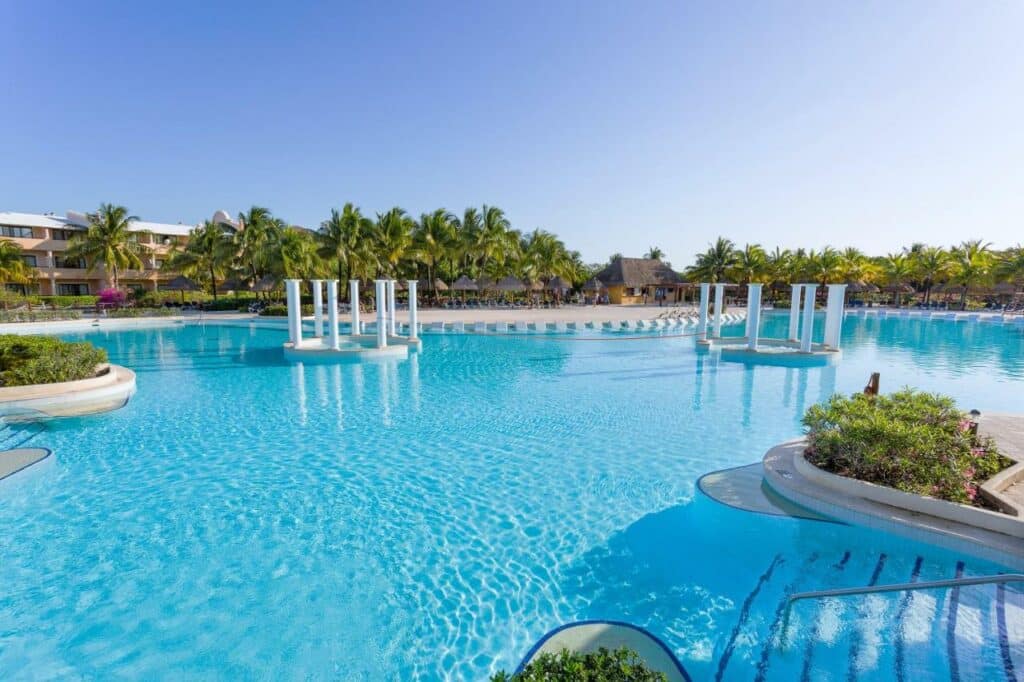 Grande piscina com água azul cristalina, cercada por palmeiras, edifícios do Grand Palladium Kantenah Resort & Spa e espreguiçadeiras ao fundo.