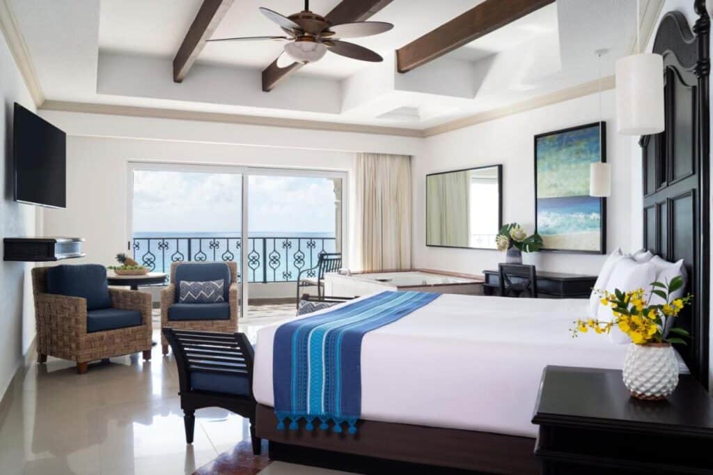 Um quarto do Hyatt Zilara Cancun dispõe de uma cama com cobertor azul, área de estar, janela grande com varanda, ventilador de teto e arte na parede.