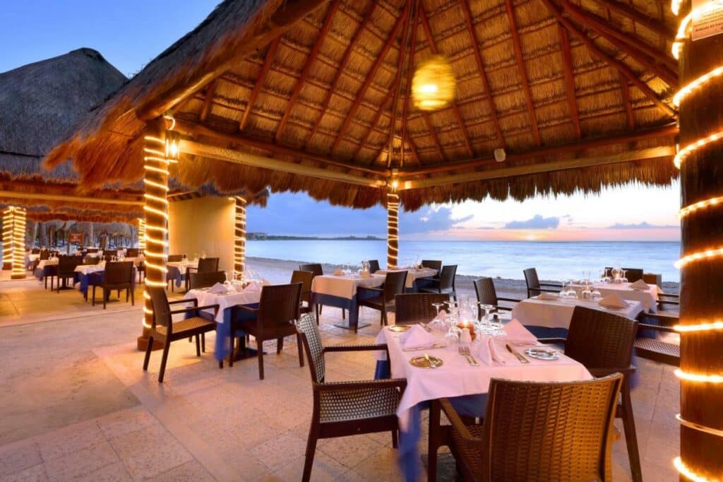 Restaurante ao ar livre no Grand Palladium Kantenah Resort & Spa à beira-mar ao pôr do sol, com telhados de palha, cadeiras de vime e mesas dispostas para refeições, envoltas em luzes de corda.