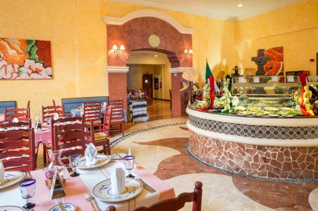 Interior aconchegante do restaurante do Grand Palladium Kantenah Resort & Spa com mesas postas, arco decorativo e uma área de bufê com vários pratos e decoração colorida. Esse é um dos melhores resorts all inclusive no Caribe.