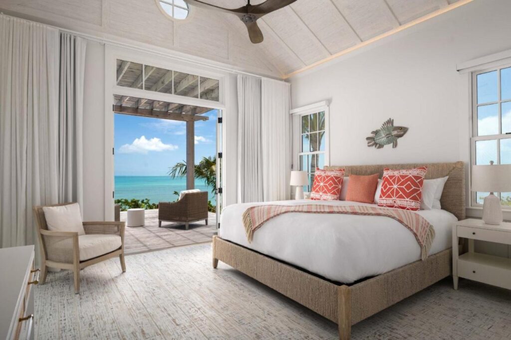 Um quarto iluminado do Ambergris Cay Private Island com uma cama, travesseiros corais e brancos, uma cadeira e vista para um pátio com vista para o oceano através de portas abertas.