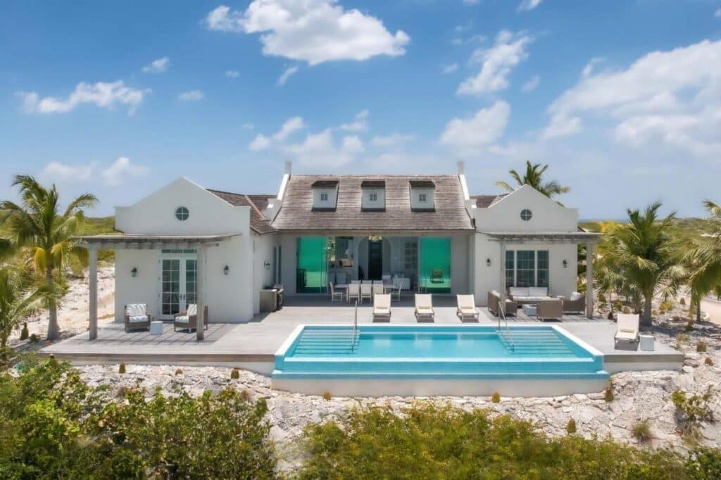 Uma casa moderna no Ambergris Cay Private Island com piscina, palmeiras e um deck sob um céu azul. Exterior branco e espreguiçadeiras são visíveis. Esse é um dos melhores resorts all inclusive no Caribe.