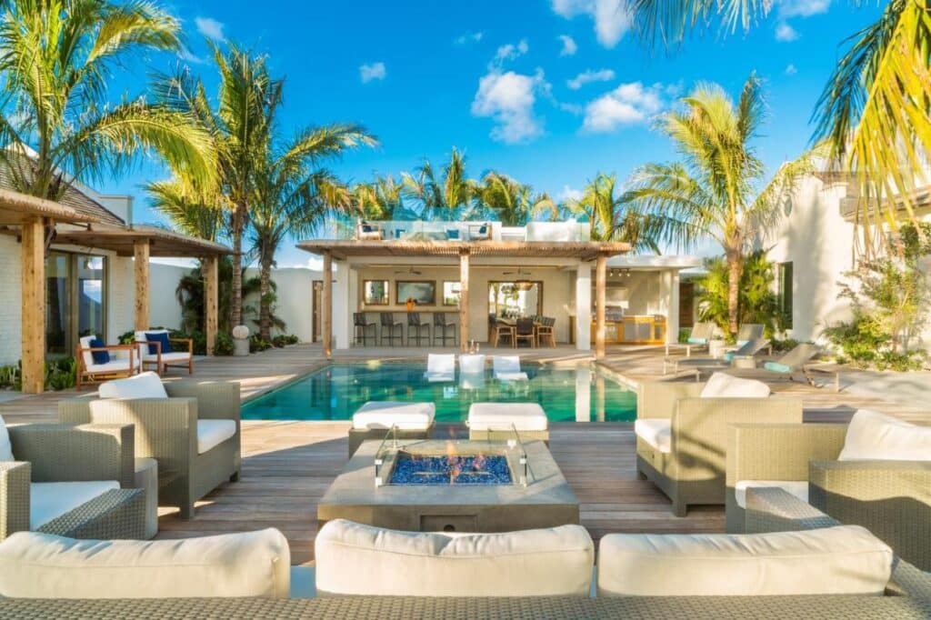 Pátio externo com móveis de vime, fogueira e piscina do Ambergris Cay Private Island. Palmeiras e um deck com área de bar sob um céu azul claro.